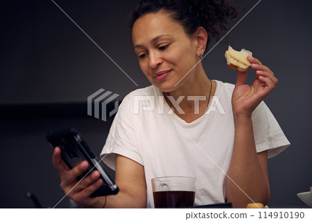 Close-up portrait pretty woman 40s, using smartphone, checking social media content, scrolling newsfeed, browsing web, standing at kitchen counter with sandwich in hand, enjoying her breakfast at home Close-up portrait pretty woman 40s, using smartphone, checking social media content, scrolling newsfeed, browsing web, standing at kitchen counter with sandwich in hand, enjoying her breakfast at home 114910190