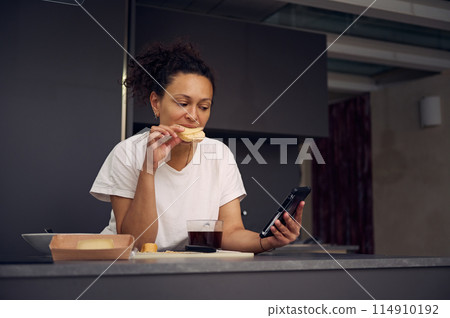 Beautiful multi ethnic woman 40s, eating sandwich with cheese and checking social media content on his smartphone, standing at kitchen counter in minimalist home kitchen interior Beautiful multi ethnic woman 40s, eating sandwich with cheese and checking social media content on his smartphone, standing at kitchen counter in minimalist home kitchen interior 114910192