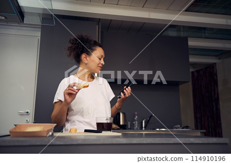Authentic multi ethnic young adult woman 40s, eating sandwich with cheese and holding her smartphone in hands, smiling looking aside, standing at kitchen counter in minimalist home kitchen interior. 114910196