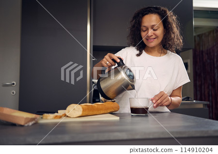 Smiling multi ethnic woman at home, making coffee. Young pretty brunette n white pajamas, standing at kitchen table, preparing breakfast, poring water from a teapot into a glass and making sandwiches Smiling multi ethnic woman at home, making coffee. Young pretty brunette n white pajamas, standing at kitchen table, preparing breakfast, poring water from a teapot into a glass and making sandwiches 114910204