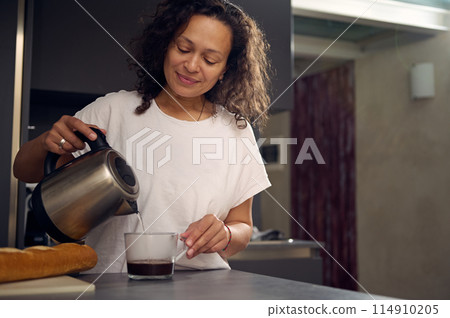 Smiling woman in white t-shirt making coffee at home, pouring boiled water from a teapot into a glass cup, standing at kitchen table with a loaf of wholesome bread on the cutting board 114910205
