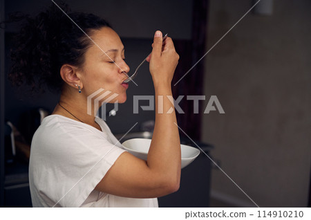 Authentic side portrait of a beautiful curly haired brunette, multi ethnic woman eating healthy muesli for breakfast in the morning. Nearby a cup of freshly brewed coffee on the kitchen counter 114910210