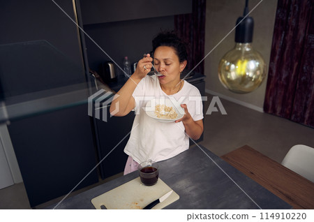 Latin American young woman 40s, smiling, drinking coffee and eating healthy muesli for breakfast in the morning at home kitchen, dressed in white pajamas Latin American young woman 40s, smiling, drinking coffee and eating healthy muesli for breakfast in the morning at home kitchen, dressed in white pajamas 114910220
