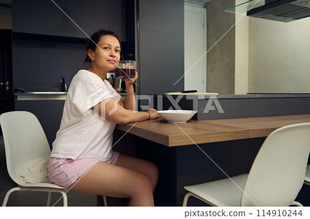 Pretty woman in pajamas, relaxing over cup of coffee. Happy young adult sitting at kitchen table, drinking hot coffee in the morning during her breakfast 114910244
