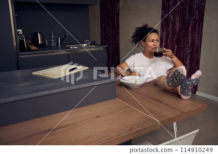 Pretty woman in pajamas, relaxing over cup of coffee. Happy young adult sitting at kitchen table, drinking hot coffee in the morning during her breakfast Pretty woman in pajamas, relaxing over cup of coffee. Happy young adult sitting at kitchen table, drinking hot coffee in the morning during her breakfast 114910249