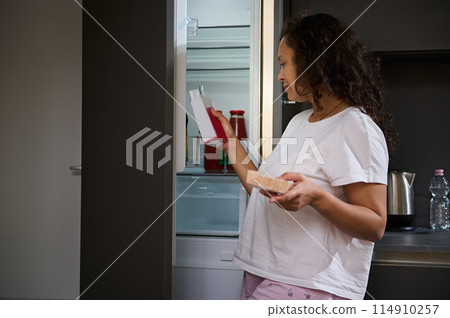 Authentic portrait of a beautiful woman standing by an open refrigerator, looking at food packaging in her hands, preparing breakfast in her home kitchen Authentic portrait of a beautiful woman standing by an open refrigerator, looking at food packaging in her hands, preparing breakfast in her home kitchen 114910257