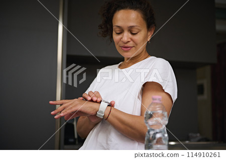 Beautiful smiling young woman checking her wearable smart wrist watch, standing at kitchen counter in the morning. A bottle of water on the foreground Beautiful smiling young woman checking her wearable smart wrist watch, standing at kitchen counter in the morning. A bottle of water on the foreground 114910261