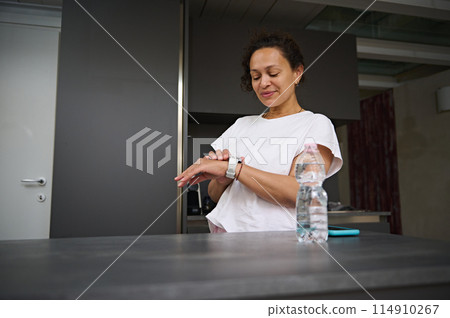 Multi ethnic smiling heathy young woman checking smart wrist watch. A bottle of pure water on the foreground. Fitness app, mobile app. People. Health care and modern wireless technology Multi ethnic smiling heathy young woman checking smart wrist watch. A bottle of pure water on the foreground. Fitness app, mobile app. People. Health care and modern wireless technology 114910267