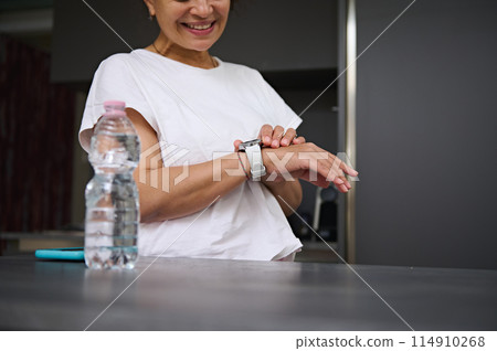Close-up view of hands of a smiling heathy young woman checking smart wrist watch. A bottle of pure water on the foreground. Fitness app, mobile app. People. Health care and modern wireless technology 114910268