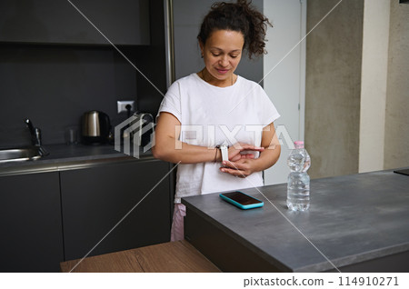 Smiling adult woman in white t-shirt putting on, wearing white smart wrist watch with black mockup digital screen, standing at kitchen counter in the minimalist home interior. Copy advertising space 114910271