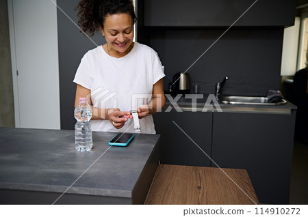 Smiling pretty woman in white t-shirt holding modern white smart wrist watch with black mockup digital screen, standing at kitchen counter in the minimalist home interior. Copy advertising space Smiling pretty woman in white t-shirt holding modern white smart wrist watch with black mockup digital screen, standing at kitchen counter in the minimalist home interior. Copy advertising space 114910272