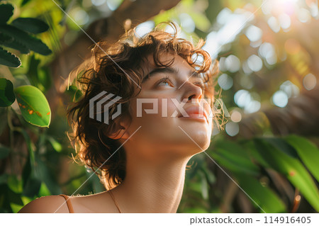 In the forest, a woman with curly hair gazes up at the sun, surrounded by branches and trees. Her eyelashes flutter in the light, happy in natures embrace In the forest, a woman with curly hair gazes up at the sun, surrounded by branches and trees. Her eyelashes flutter in the light, happy in natures embrace 114916405