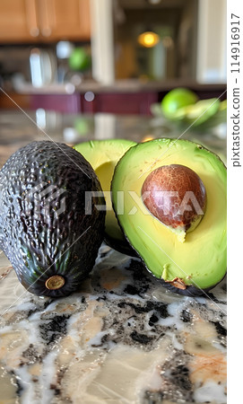 Two avocados, natural foods and fruits, are sitting on a counter. One avocado is cut in half, revealing its superfood qualities as a nutritious ingredient 114916917