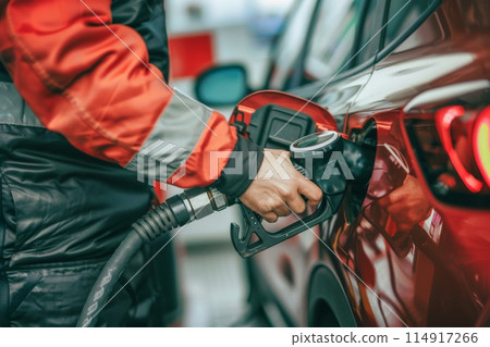 At a gas station, a person in a yellow jacket is fueling up a car using a fuel nozzle At a gas station, a person in a yellow jacket is fueling up a car using a fuel nozzle 114917266