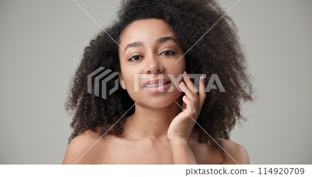 Beauty and healthcare concept - beautiful African American woman with curly afro hairstyle and clean, healthy skin touching face with hand, doing facial massage, posing and looking at camera Beauty and healthcare concept - beautiful African American woman with curly afro hairstyle and clean, healthy skin touching face with hand, doing facial massage, posing and looking at camera 114920709