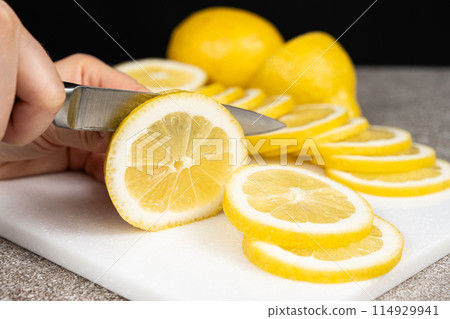 Woman cutting lemon into slices with a sharp utility knife on a stone kitchen board. Woman cutting lemon into slices with a sharp utility knife on a stone kitchen board. 114929941