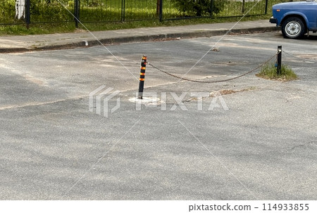 Metal restraints with an iron chain with a key for parking a car between poles outdoors in a special area on a summer day. There is an unknown car next to them Metal restraints with an iron chain with a key for parking a car between poles outdoors in a special area on a summer day. There is an unknown car next to them 114933855