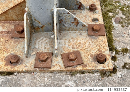 Metal pillar fastened to basement with anchor bolts and nuts. The rusty surface of the post and the fastenings are in places. The support of the iron pillar on a green lawn in the park. Close-up 114933887