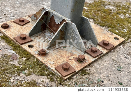 Metal pillar fastened to basement with anchor bolts and nuts. The rusty surface of the post and the fastenings are in places. The support of the iron pillar on a green lawn in the park. Close-up 114933888