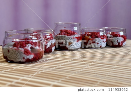 A lot of healthy Chia pudding with coconut milk in a glass containers in view can with fresh berries as raspberry, red currant, cranberry and pieces of peach on bamboo background. Close-up A lot of healthy Chia pudding with coconut milk in a glass containers in view can with fresh berries as raspberry, red currant, cranberry and pieces of peach on bamboo background. Close-up 114933932