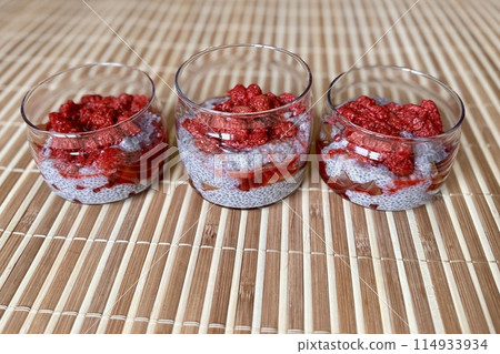 A lot of healthy Chia pudding with coconut milk in a glass containers in view can with fresh berries as raspberry, red currant, cranberry and pieces of peach on bamboo background. Close-up A lot of healthy Chia pudding with coconut milk in a glass containers in view can with fresh berries as raspberry, red currant, cranberry and pieces of peach on bamboo background. Close-up 114933934