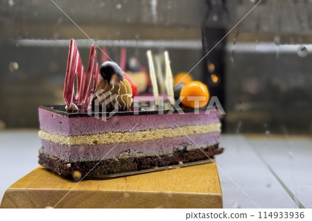 A portion of layered cream chocolate cake with blueberries, blackberries, sea buckthorn on a wooden stand on the counter of the restaurant. Close-up A portion of layered cream chocolate cake with blueberries, blackberries, sea buckthorn on a wooden stand on the counter of the restaurant. Close-up 114933936