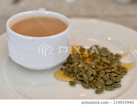 A cup of coffee sits beside a sound of pumpkin seeds poured with liquid lime honey, showcasing a contract between the liquid beverage and the solid ingredient at the table. Close-up A cup of coffee sits beside a sound of pumpkin seeds poured with liquid lime honey, showcasing a contract between the liquid beverage and the solid ingredient at the table. Close-up 114933940