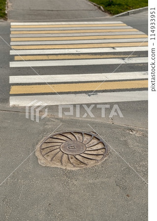 A sunny day view of a crosswalk with alternating yellow and white stripes beside a manhole cover A sunny day view of a crosswalk with alternating yellow and white stripes beside a manhole cover 114933951
