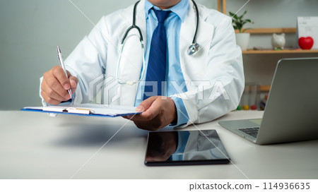 Male doctor sitting at work desk checking work plans and writing document reports in hospital office, healthcare worker and doctor service, digital laptop in modern office, medical technology concept. 114936635