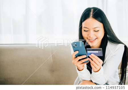 Smiling woman with smartphone and credit card shops online on sofa. Engaged in ordering banking and buying goods or services. Modern technology for convenient shopping at home. 114937105