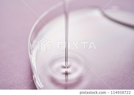Serum or cosmetic oil flows into a transparent bowl on a purple background. 114938722