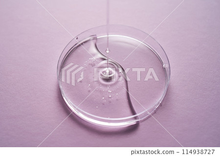 Serum or cosmetic oil flows into a transparent bowl on a purple background. 114938727