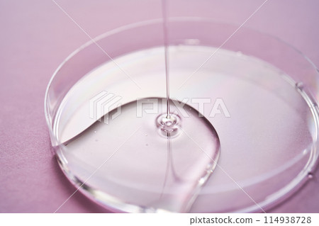 Serum or cosmetic oil flows into a transparent bowl on a purple background. Serum or cosmetic oil flows into a transparent bowl on a purple background. 114938728
