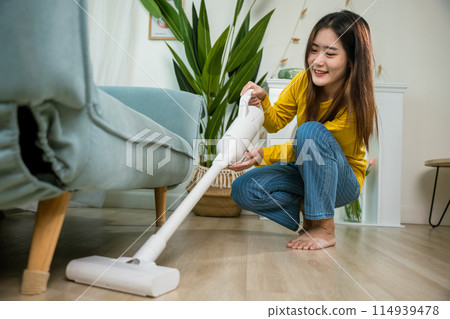Housewife female dust cleaning floor under sofa or couch furniture with vacuum cleaner, Happy Asian young woman with accumulator vacuum cleaner at home in living room, household and housework concept 114939478