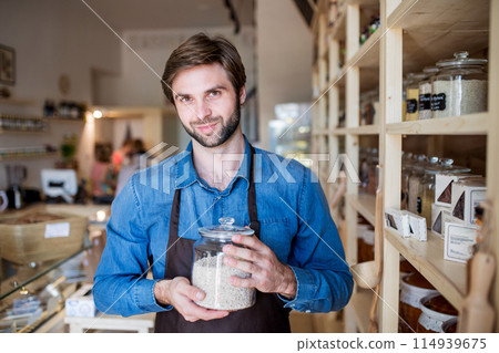 Handsome man working in package-free store using reusable containers. Zero waste shops offering package-free bulk goods and sustainable alternatives. Handsome man working in package-free store using reusable containers. Zero waste shops offering package-free bulk goods and sustainable alternatives. 114939675