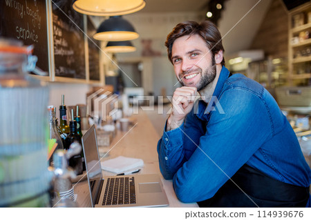 Man in small business, working in his bistro, writing email to supplier. Young male business owner. Man in small business, working in his bistro, writing email to supplier. Young male business owner. 114939676