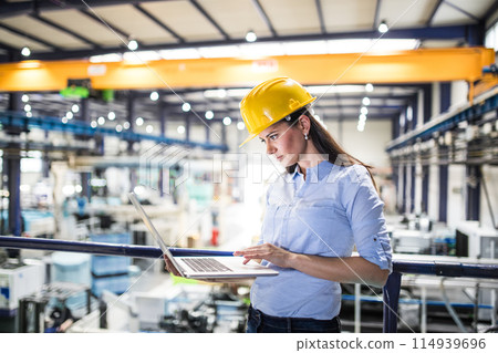 Female project manager standing in modern industrial factory. Manufacturing facility with robotics, robotic arms and automation. Storing products and materials in warehouse. 114939696