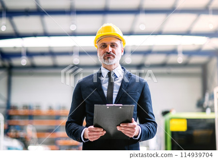 Portrait of project manager standing in modern industrial factory. Manufacturing facility with robotics, robotic arms and automation. Storing products and materials in warehouse. 114939704
