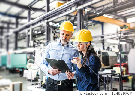 Two project managers standing in modern industrial factory. Manufacturing facility with robotics, robotic arms and automation. Storing products and materials in warehouse. 114939707
