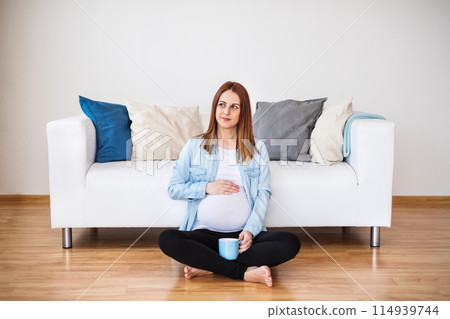 Pregnant woman sitting on floor in front of sofa, drinking herbal tea. Red raspberry leaf tea to induce labor. 114939744