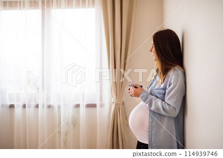 Pregnant woman leaning against wall in her home, drinking herbal tea. Red raspberry leaf tea to induce labor. 114939746