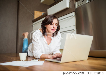 Mature woman working from homeoffice. Woman paying bills online, lying on floor. Mature woman working from homeoffice. Woman paying bills online, lying on floor. 114939848
