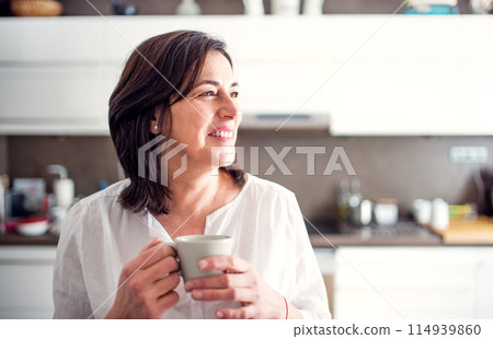 Beautiful mature woman in kitchen, drinking coffee. Older woman living alone, enjoying peaceful weekend day Beautiful mature woman in kitchen, drinking coffee. Older woman living alone, enjoying peaceful weekend day 114939860