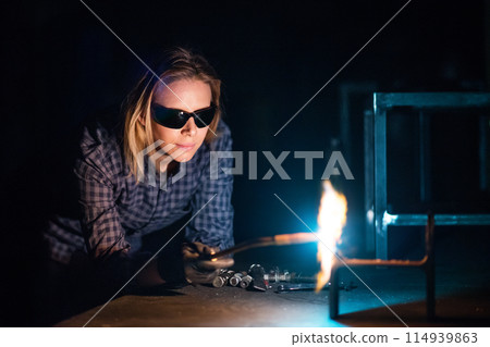 Beautiful blonde woman works as a welder in workshop, operating welding machine, wearing safety goggles for eye protection. 114939863