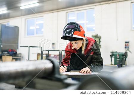 Beautiful blonde woman works as a welder in workshop, operating welding machine, wearing protective clothing and a welding mask. 114939864