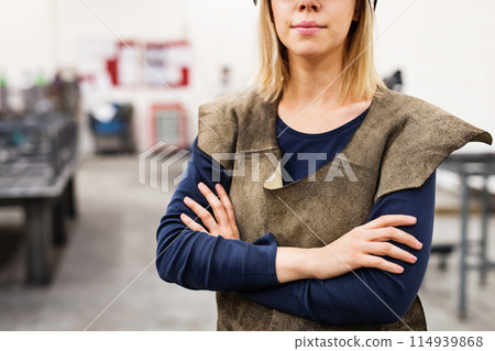 Beautiful blonde woman works as a welder in workshop, wearing leather welding apron. 114939868