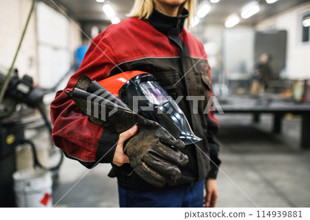 Close up of welding mask in hands of woman welder in workshop. Female worker operating welding machine, wearing protective clothing and a welding mask. 114939881