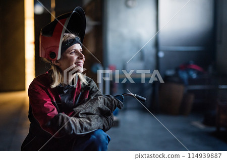 Beautiful blonde woman works as a welder in workshop, operating welding machine, wearing protective clothing and a welding mask. 114939887