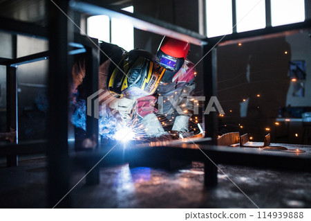 Blonde woman works as a welder in workshop, operating welding machine, wearing protective clothing and a welding mask. Two welders helpign each other. Blonde woman works as a welder in workshop, operating welding machine, wearing protective clothing and a welding mask. Two welders helpign each other. 114939888