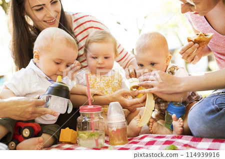 Mothers and babies enjoying group picnic outdoor in park, sitting on picnic blanket and preparing food and drinks. Mothers and babies enjoying group picnic outdoor in park, sitting on picnic blanket and preparing food and drinks. 114939916